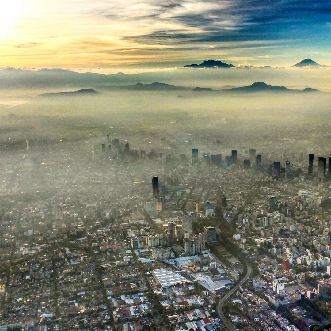 Aerial view of a city covered in smog with distant mountains visible through the haze, showing poor air quality and pollution.