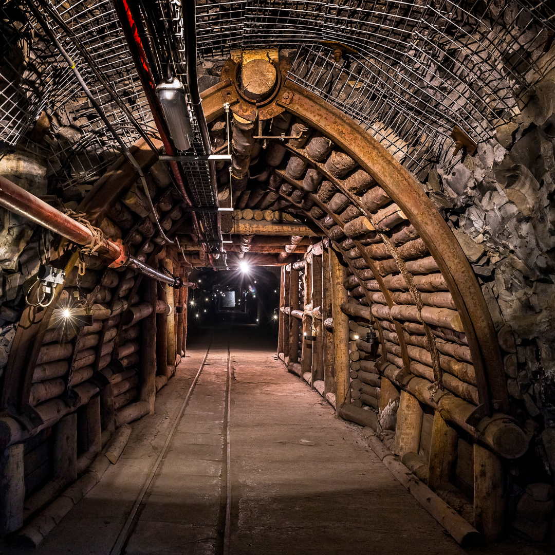 Underground mining tunnel reinforced with timber and metal supports, featuring lighting and safety equipment along the walls and ceiling.